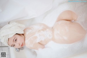 A naked asian woman standing in the shower with her hands on her head.