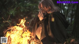 A woman sitting in front of a fire in the woods.